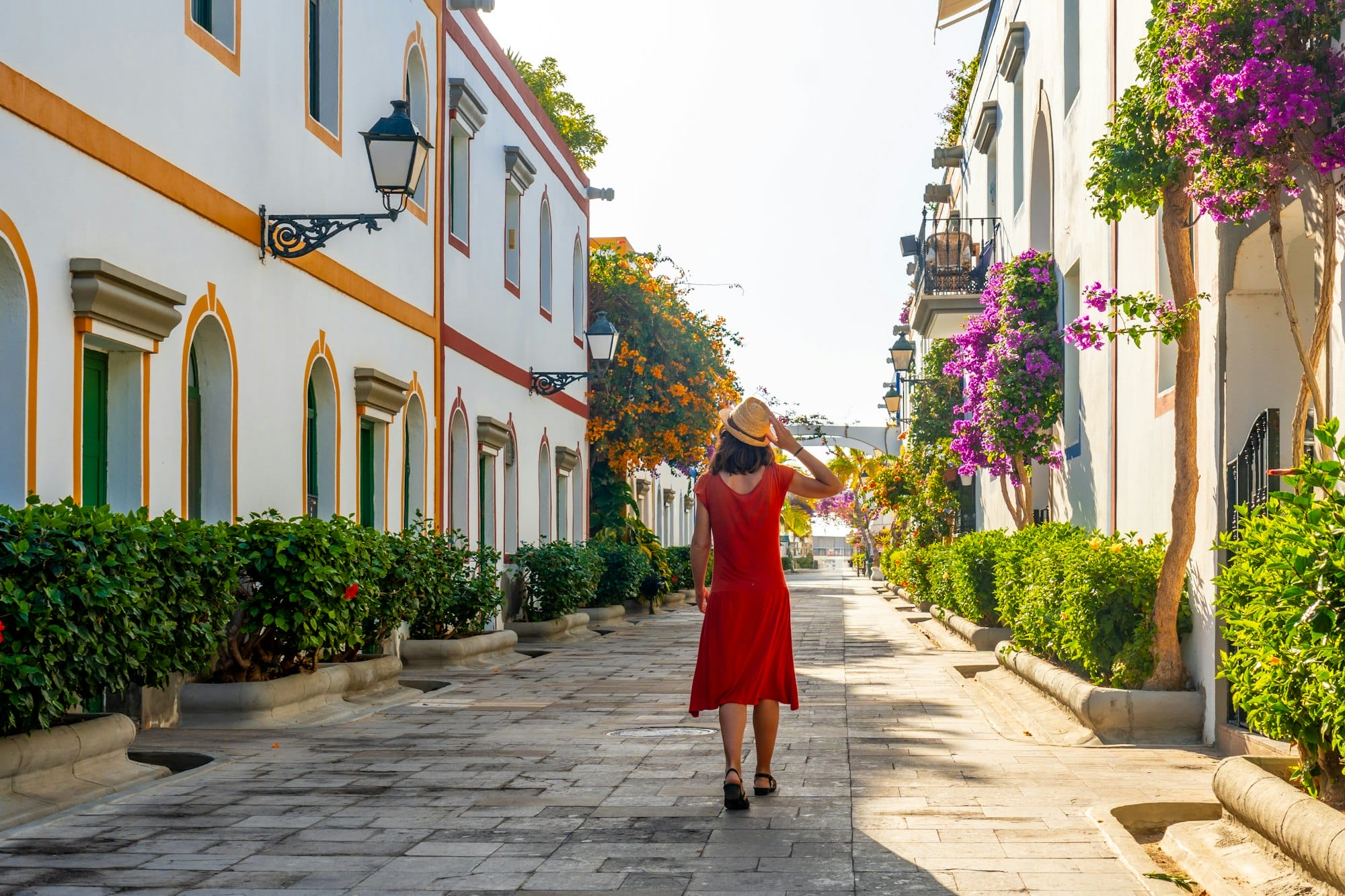 A woman in a red dress walks down a narrow street with a hat on